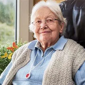 Close-up of an elderly woman who appears to be in her 70s or 80s, with white hair and glasses. She has a pleasant expression, smiling gently. She is wearing a light beige or cream-colored, knitted, or textured sweater/cardigan over a light blue collared shirt.