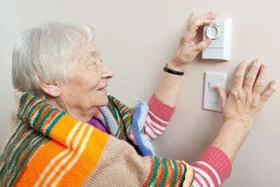 Full indoor shot of an elderly woman adjusting a thermostat and light switch on a wall.