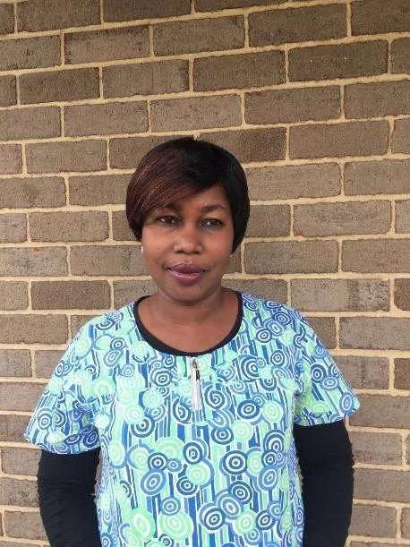 A woman with short, dark brown hair is standing in front of a light brownish-gray brick wall.