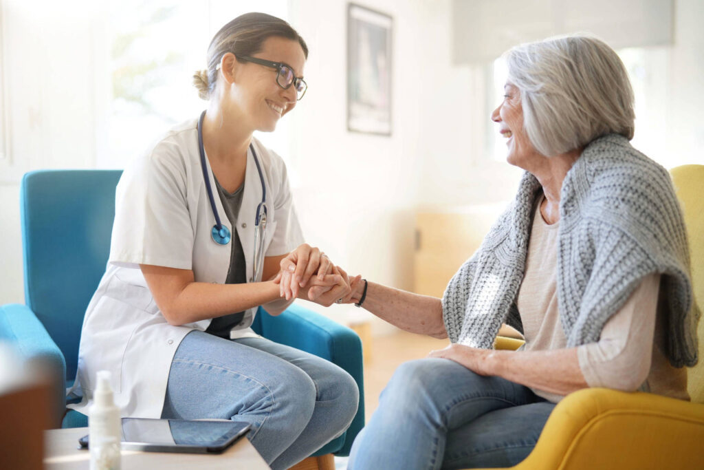Full indoor shot of a doctor holding hands with an elderly woman. The doctor, a light-skinned woman with shoulder-length brown hair pulled back, is seated in a teal-colored armchair. She is wearing a white doctor's coat over a light wash blue-colored shirt/pants. She is looking at the elderly woman. A stethoscope is visible around her neck. She is holding the elderly woman's hands. The elderly woman, light-skinned with short gray hair, is seated in a mustard-yellow armchair.