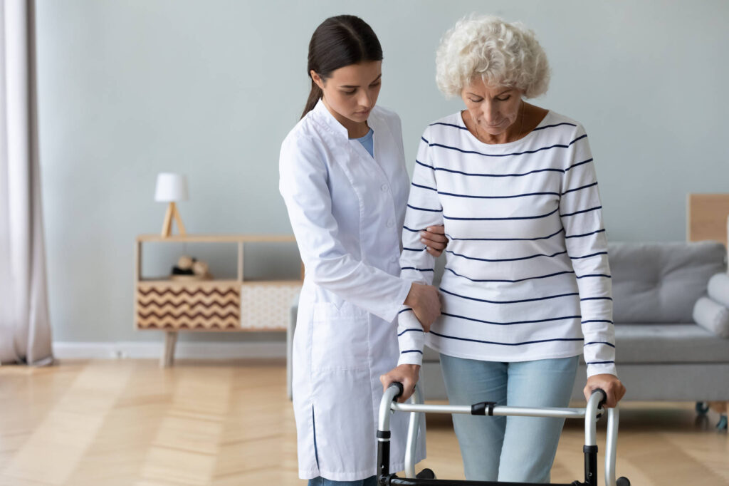 Full indoor shot of a young woman in a white lab coat assisting an older woman with walking.