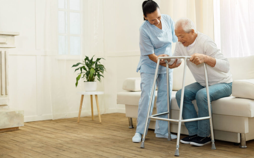 Full indoor shot of a caregiver assisting an elderly man to use a walker.