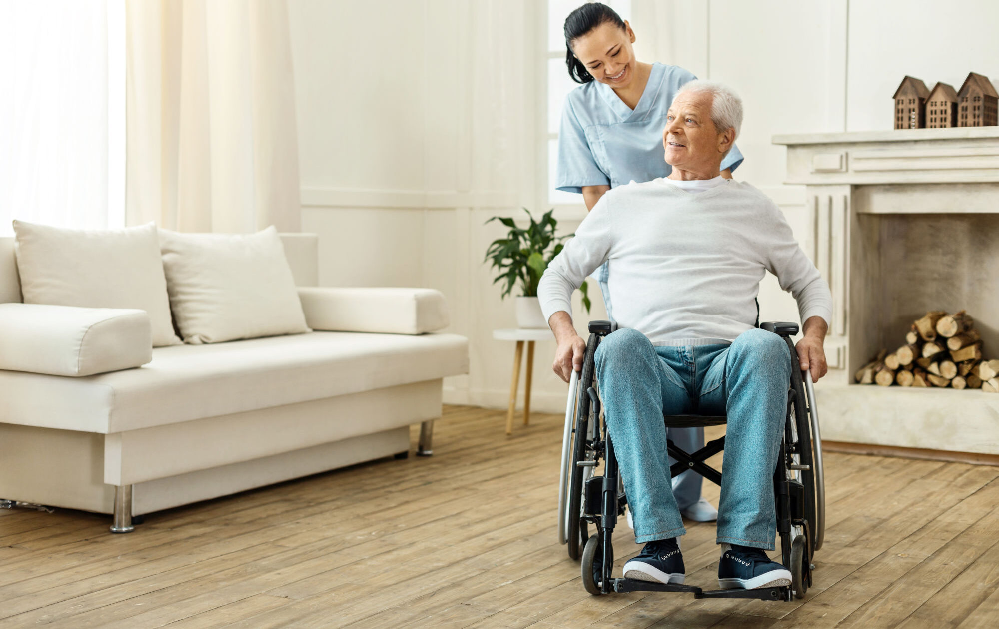Full indoor shot of a smiling woman in a light-blue scrubs assisting a man in a wheelchair.