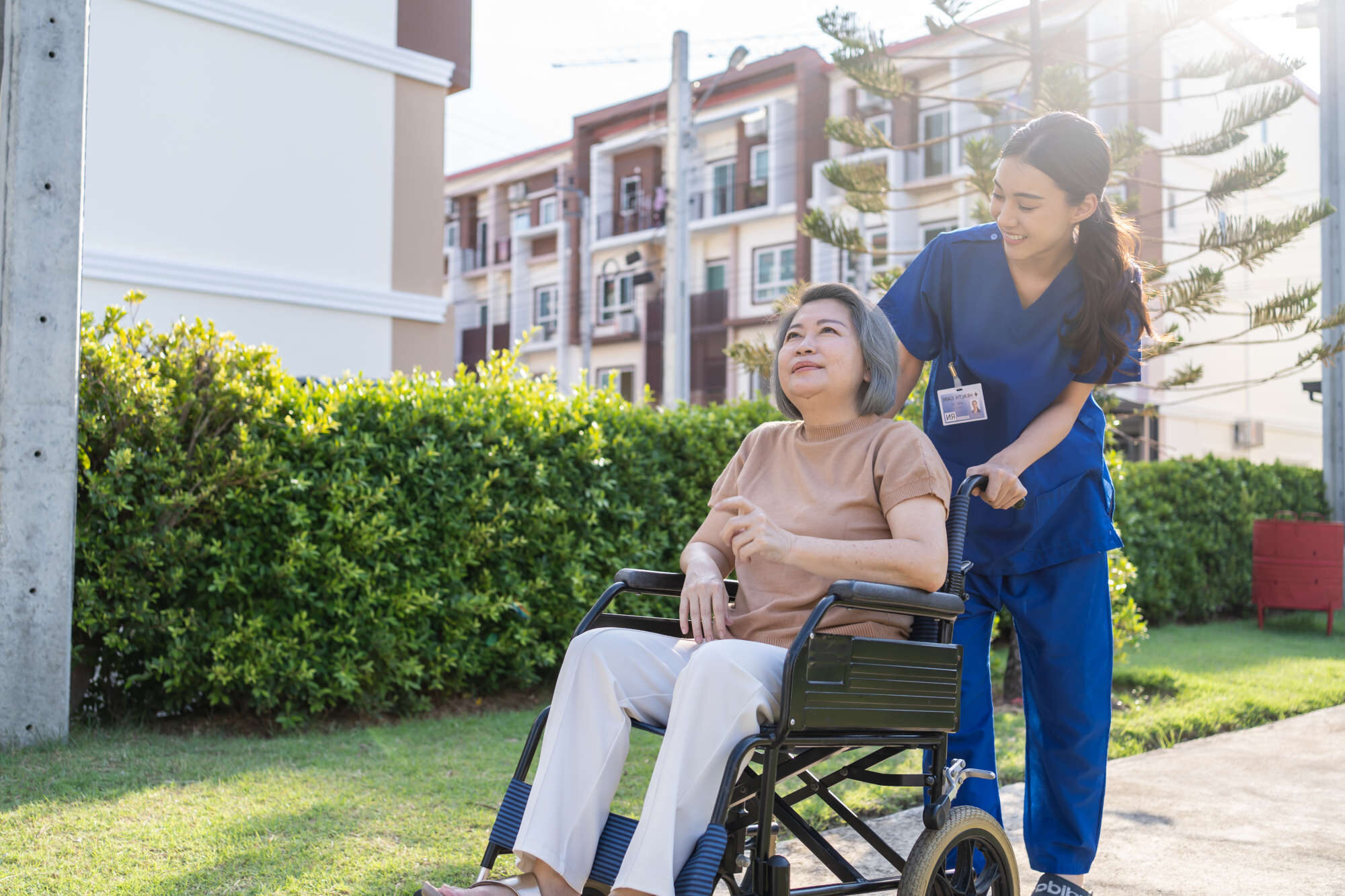 Full outdoor shot of a caregiver helping an elderly woman in a wheelchair.