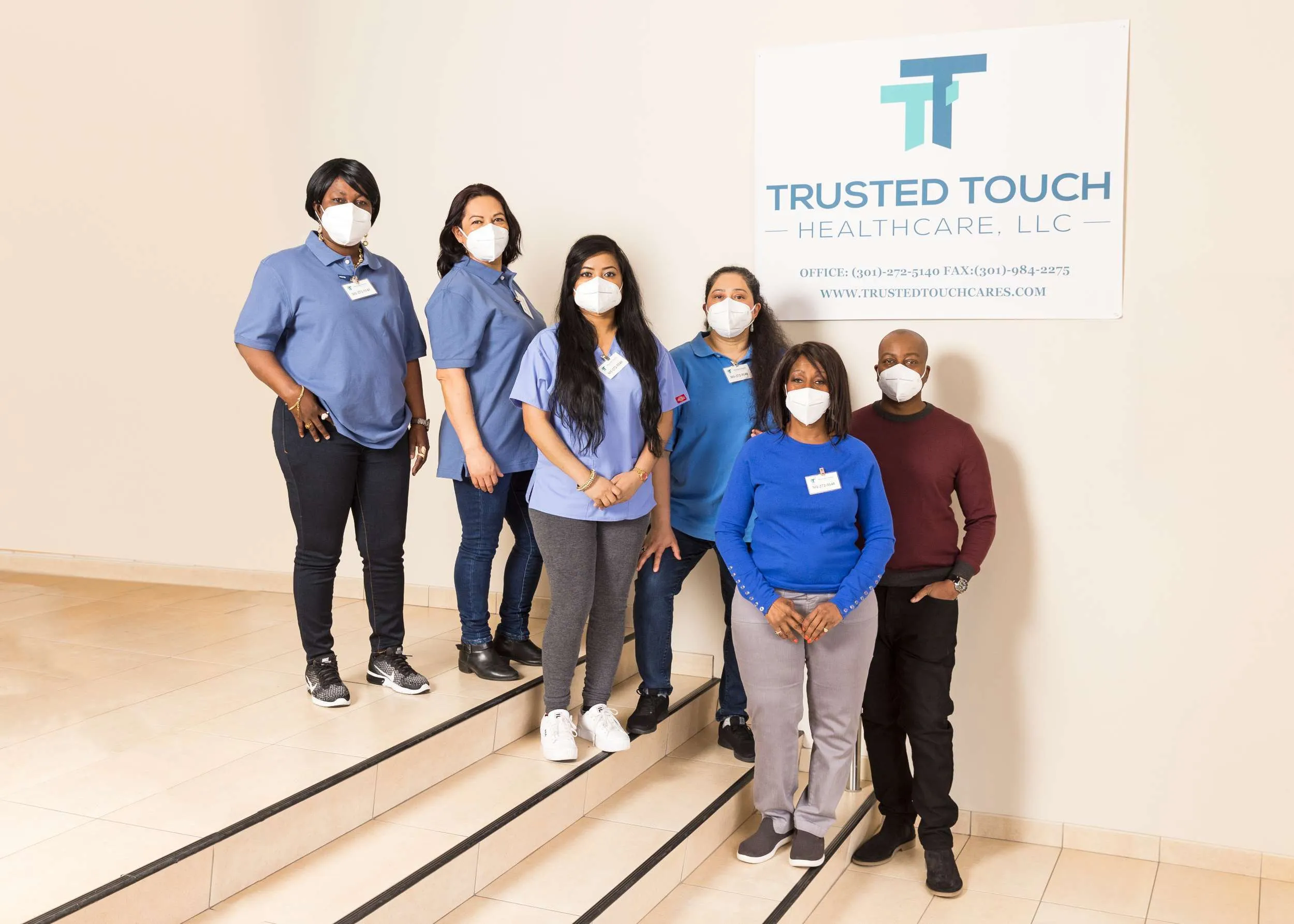 Full shot of a group of healthcare workers, standing on a staircase in front of a wall. A banner is hanging on the wall behind them that displays the name "TRUSTED TOUCH HEALTHCARE, LLC" and contact information. The individuals are wearing various colors of clothing, including blue, gray, and maroon tops.