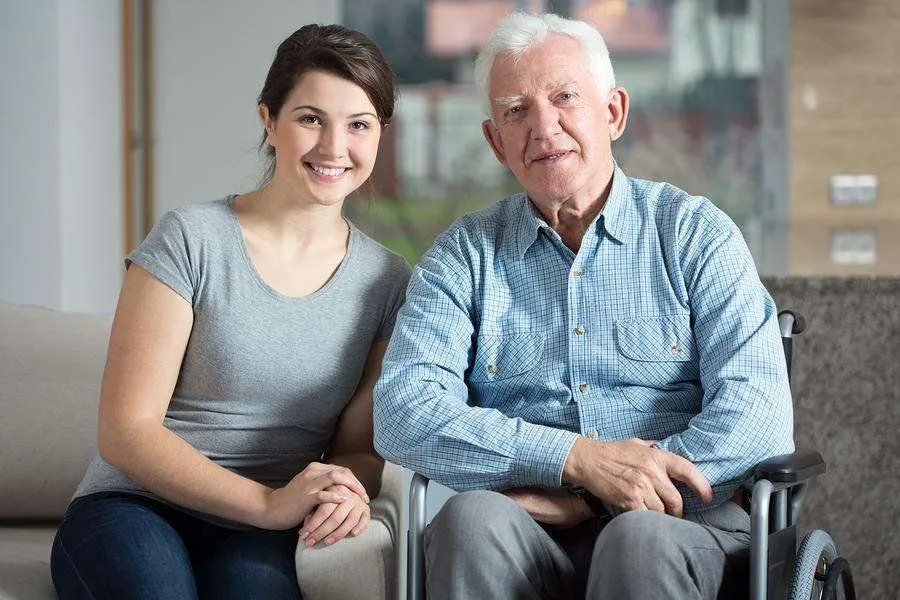 A young woman and an older man are seated. The woman, on the left side is wearing a gray t-shirt and dark-wash jeans. She is smiling and looking directly at the camera. Her hair is light brown and pulled back. The man, on the right side is wearing a light blue, button-down shirt. He is also smiling and looking directly at the camera. He is seated in a wheelchair.