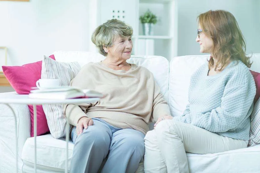 Two senior women are seated on a white couch, engaged in conversation.