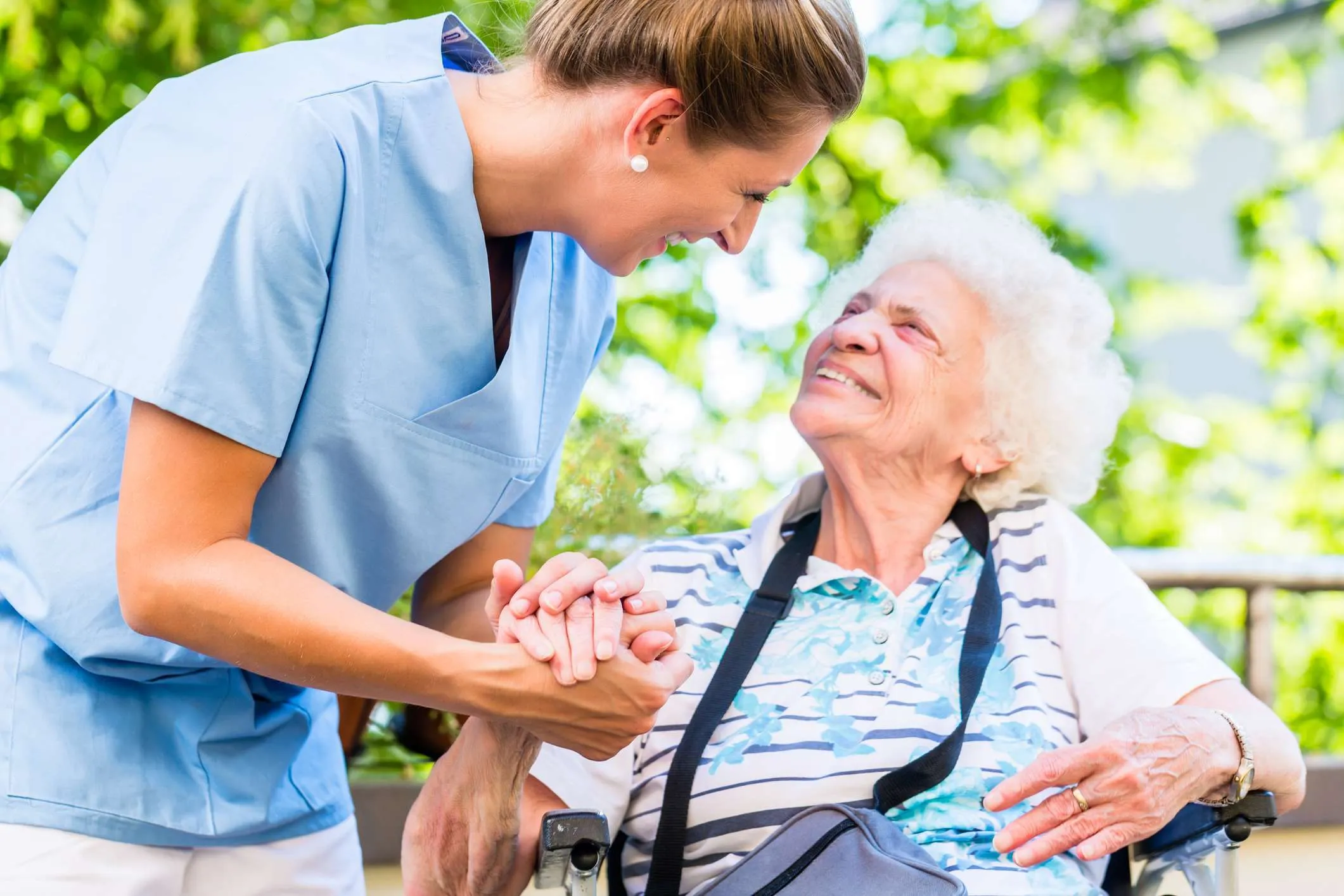 Close-up of a young woman in a light-blue medical uniform, and an elderly woman in a wheelchair. The young woman is bending down, holding the hands of the elderly woman. Her face is directed towards the elderly woman, and she is smiling gently.