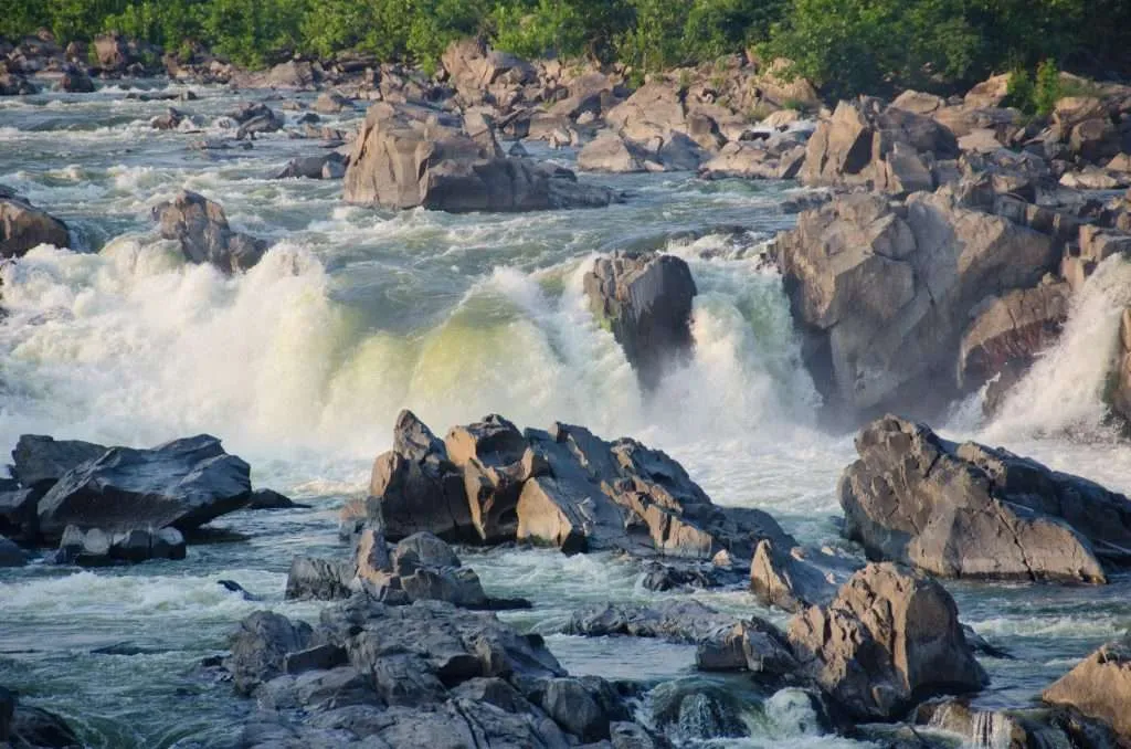 Full outdoor shot of the Great Falls of the Potomac River. The foreground showcases a rocky riverbed, with large, dark gray and brown boulders of varying sizes scattered across the river bottom.