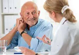 A medium shot of a man and a woman, likely in a medical setting. The man, who appears to be elderly, is seated and looking slightly towards the woman. The woman, who is likely a medical professional, is positioned slightly behind and to the right of the man.