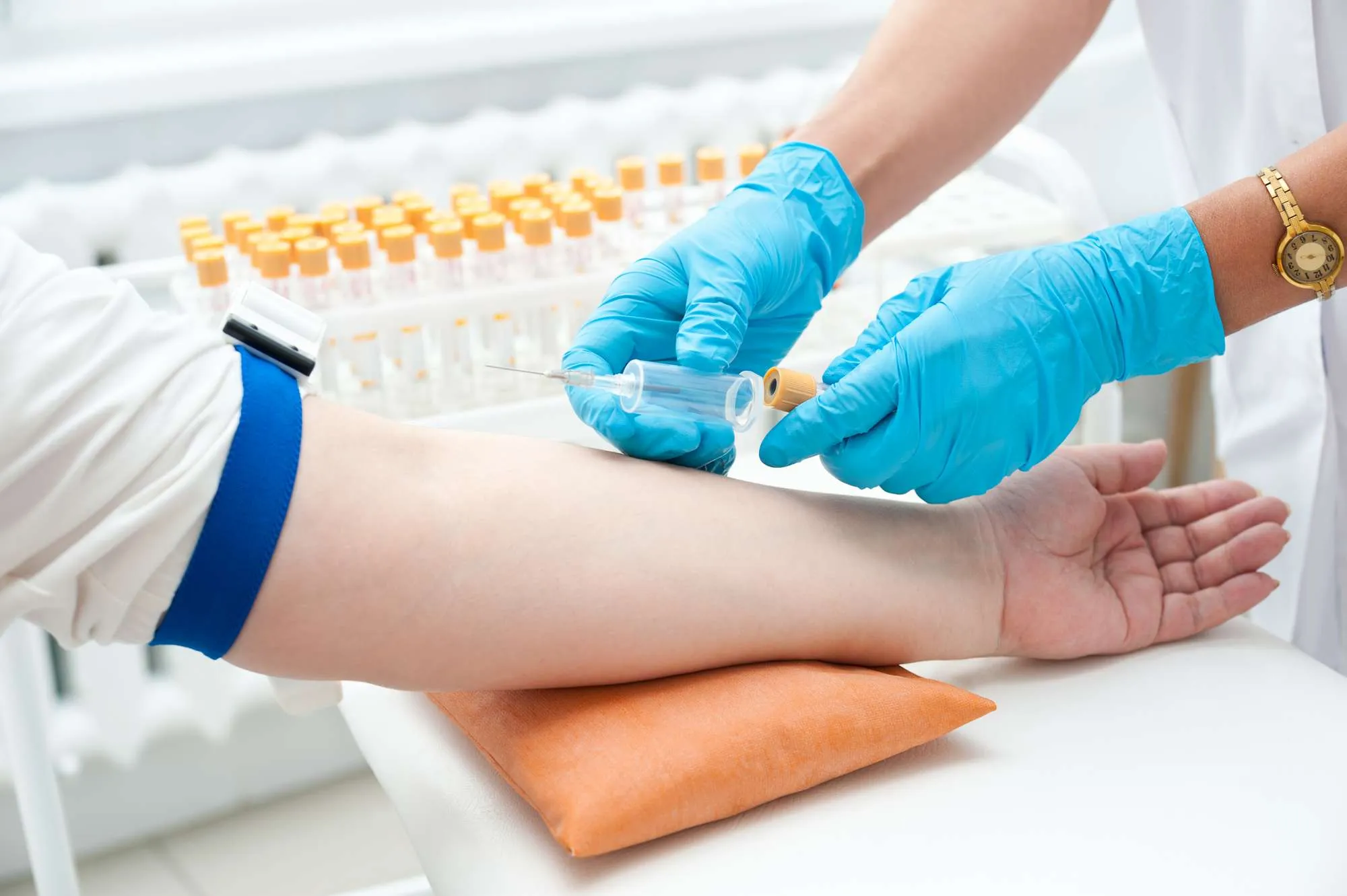 A person's arm, with a blue elastic band around the upper arm, is positioned on a table or examining surface. A medical professional, wearing light-colored medical-grade clothing and bright-blue disposable gloves, is drawing blood from the patient's arm.