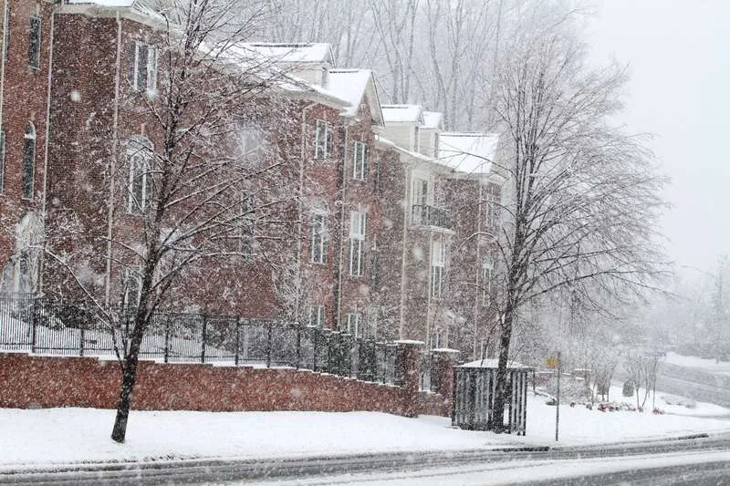 Full outdoor shot of a snowy residential area. Several brick townhouses or apartment buildings are visible, with snow covering the roofs and ground. The brickwork of the buildings is a light reddish-brown color. The snow appears to be falling softly and lightly accumulating on the ground and on the trees and buildings.