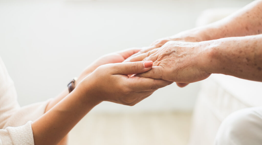 Close-up of two hands clasped together. The hands of a younger person, light-skinned with visible veins, are gently cradling the hands of an older person, also light-skinned but with noticeable wrinkles and age spots.