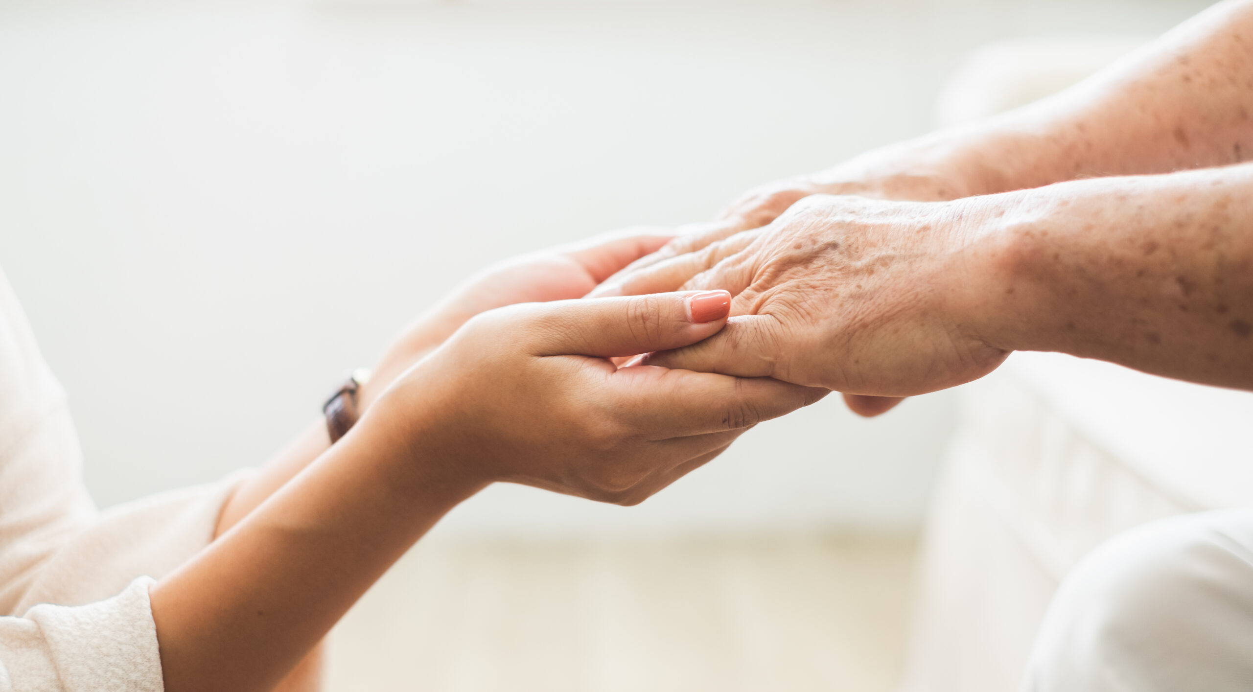 Close-up of two hands clasped together. The hands of a younger person, light-skinned with visible veins, are gently cradling the hands of an older person, also light-skinned but with noticeable wrinkles and age spots.
